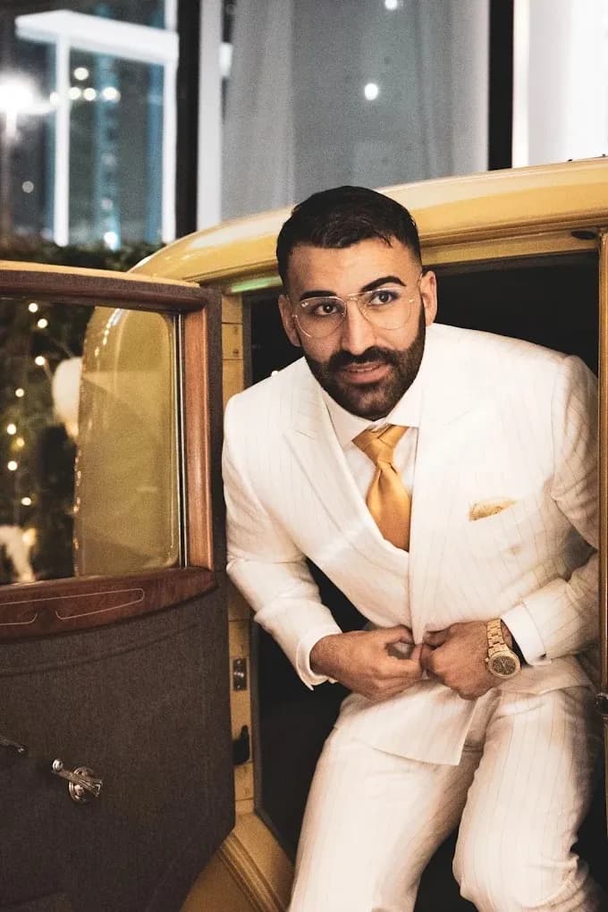 Man in an ivory pinstripe three-piece suit with a gold tie leaning out of a yellow vintage car