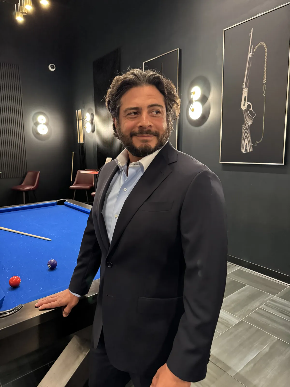 Man wearing a charcoal suit and pale blue open-collar shirt beside a blue pool table in a dark lounge