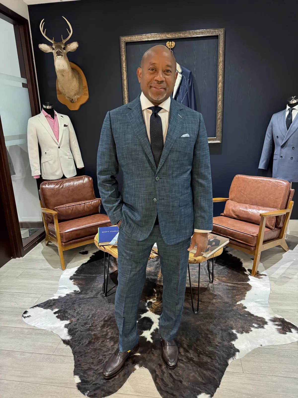 Man in a dark teal textured suit with a white shirt, navy dotted tie and light blue pocket square in an upscale menswear boutique