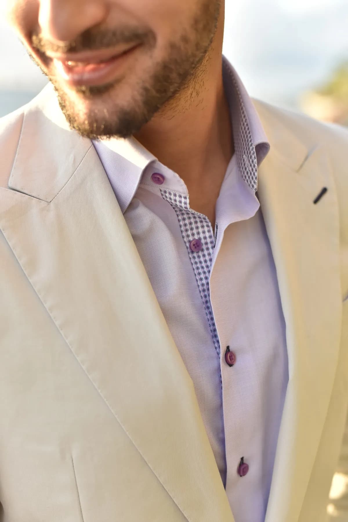 Close-up of a man wearing a cream blazer over a pale lavender open-collar shirt with patterned trim and purple buttons