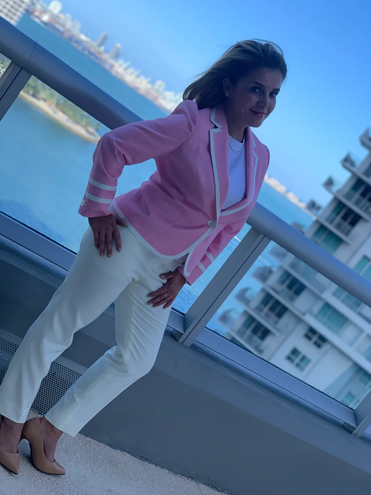 Woman in pink blazer with white piping and white trousers on waterfront balcony with city skyline