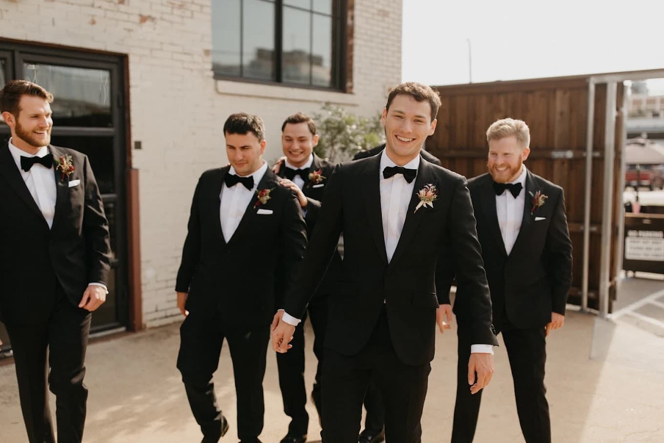 Groomsmen in matching black tuxedos with boutonnieres walking together by Bespoke By CB Miami
