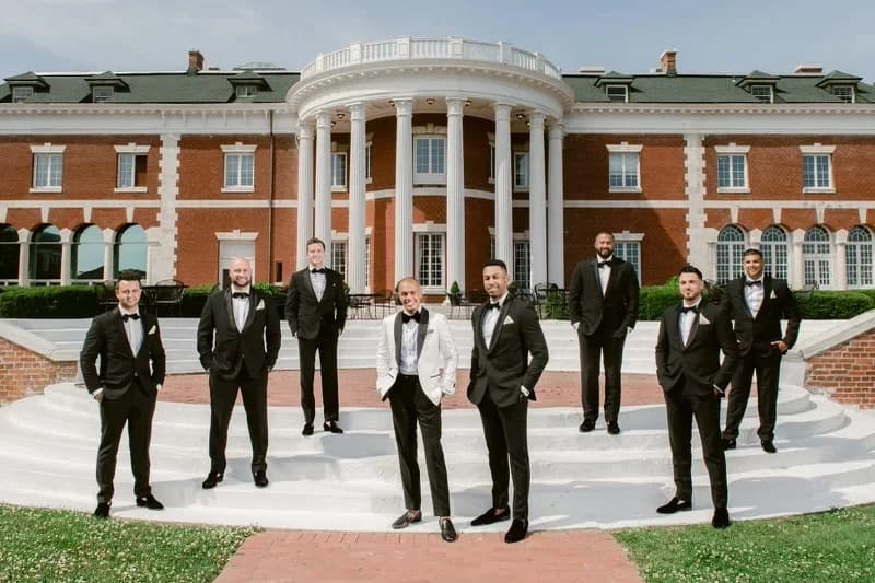 Groom in white tuxedo jacket with groomsmen in black tuxedos outside brick building by Bespoke By CB Miami