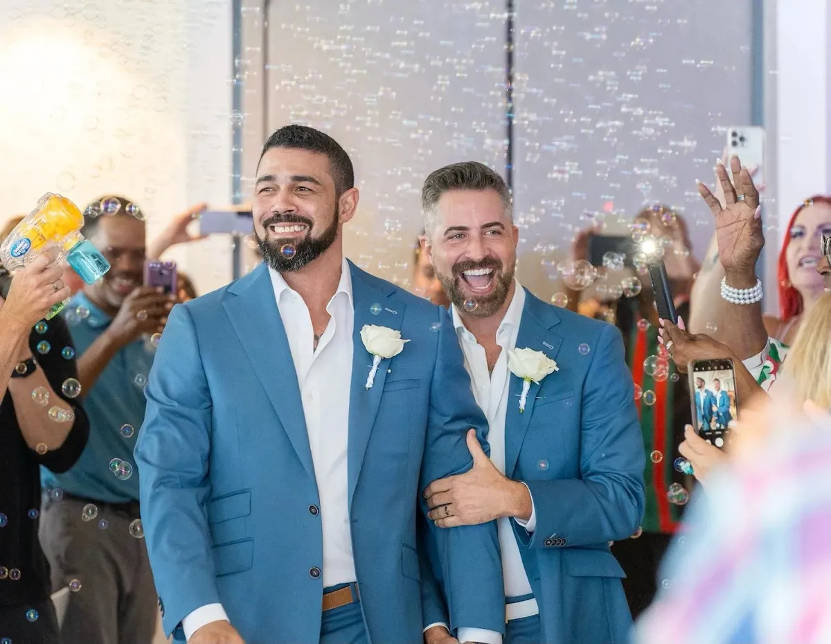 Two grooms in blue suits walking through bubble exit at wedding recessional