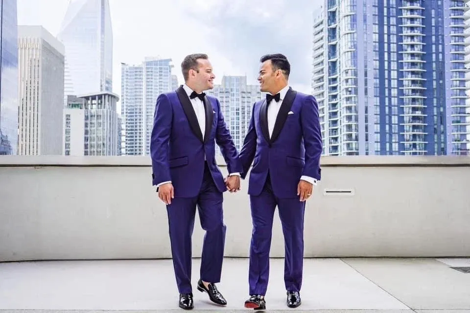 Two grooms in royal blue tuxedos holding hands on rooftop terrace with city skyline