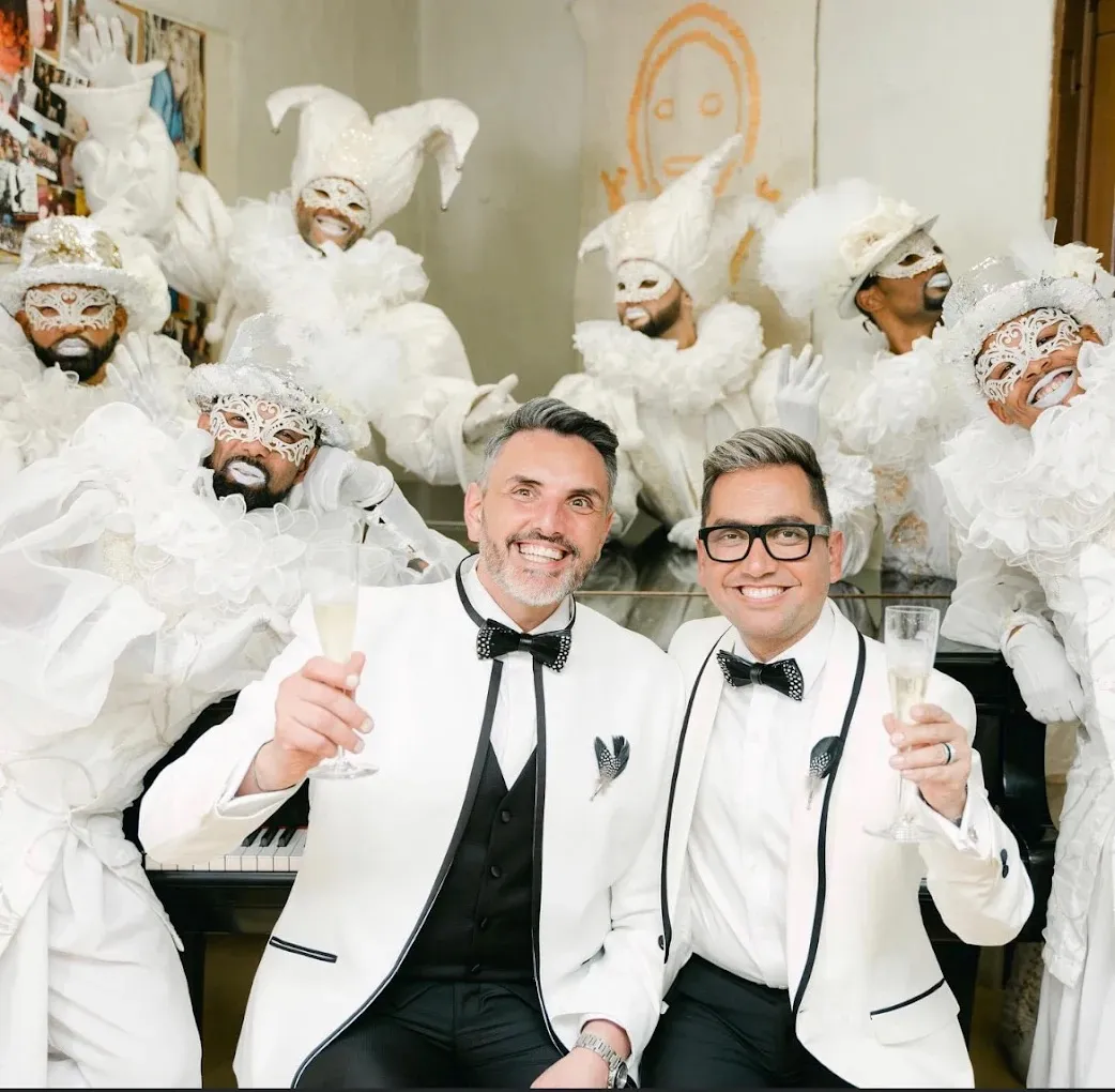 Two grooms in white tuxedo jackets toasting champagne at wedding celebration by Bespoke By CB Miami