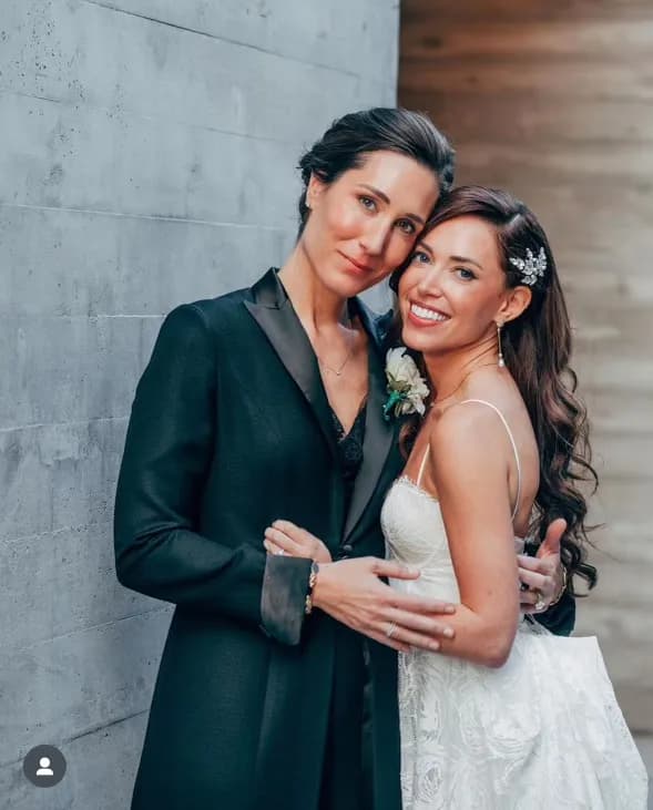 Two brides embracing, one in black wedding suit and one in white lace gown